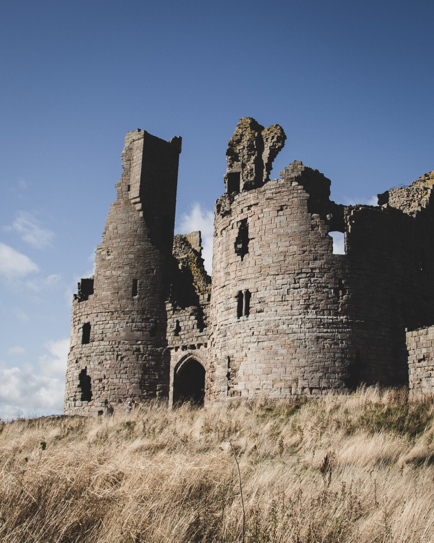 A closer view of Dunstanburgh Castle's crumbling towers. Dry, yellow grass in the foreground. 