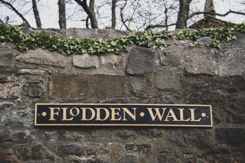 A sign for the flodden wall - a defensive structure built after the Battle of Flodden in 1513. An ivy plant trails over the top of the wall. 