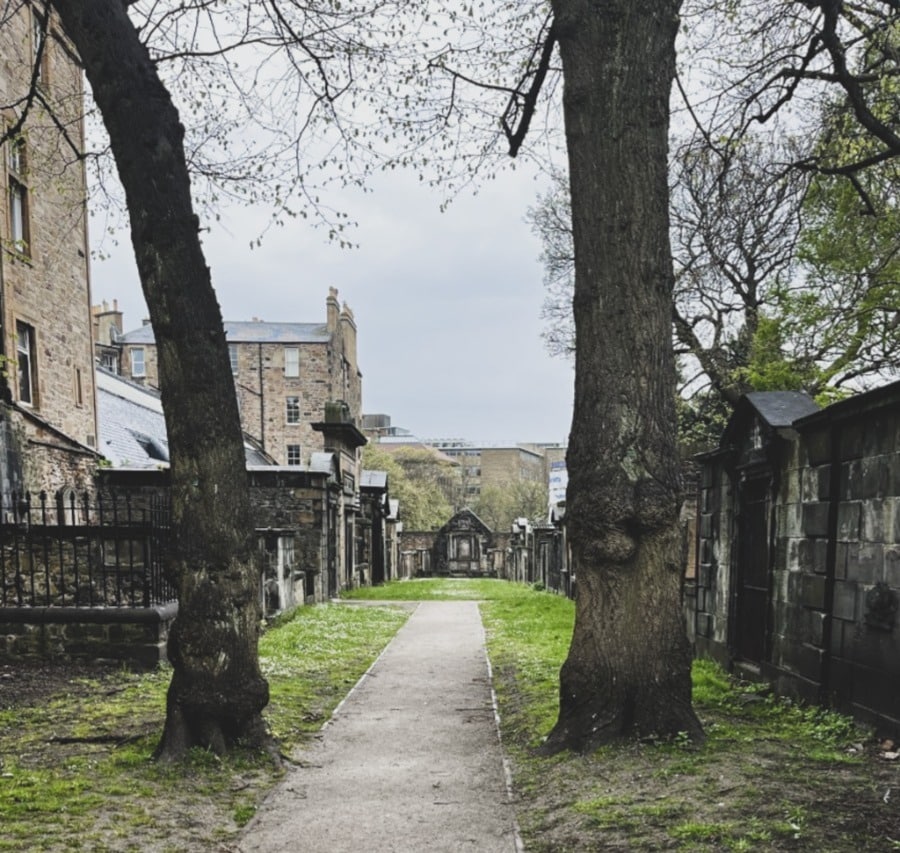 A glimpse into the Covenanters Prison. It's now an outdoor area. A path stretches into the distance. There are tombs and headstones on either side. Two trees are growing in the foreground, 